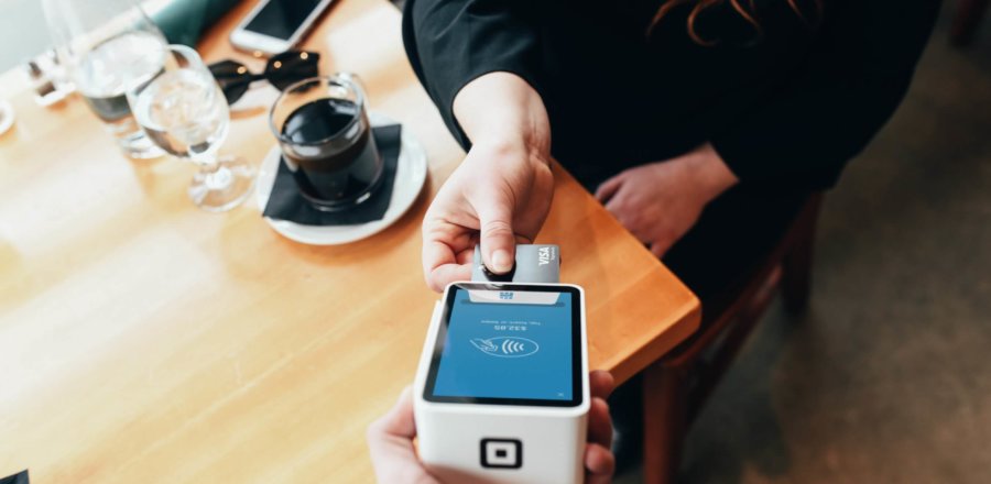 ChatGPT: Person bezahlt kontaktlos mit Karte an einem mobilen Lesegerät in einem Café mit Holz­tisch.