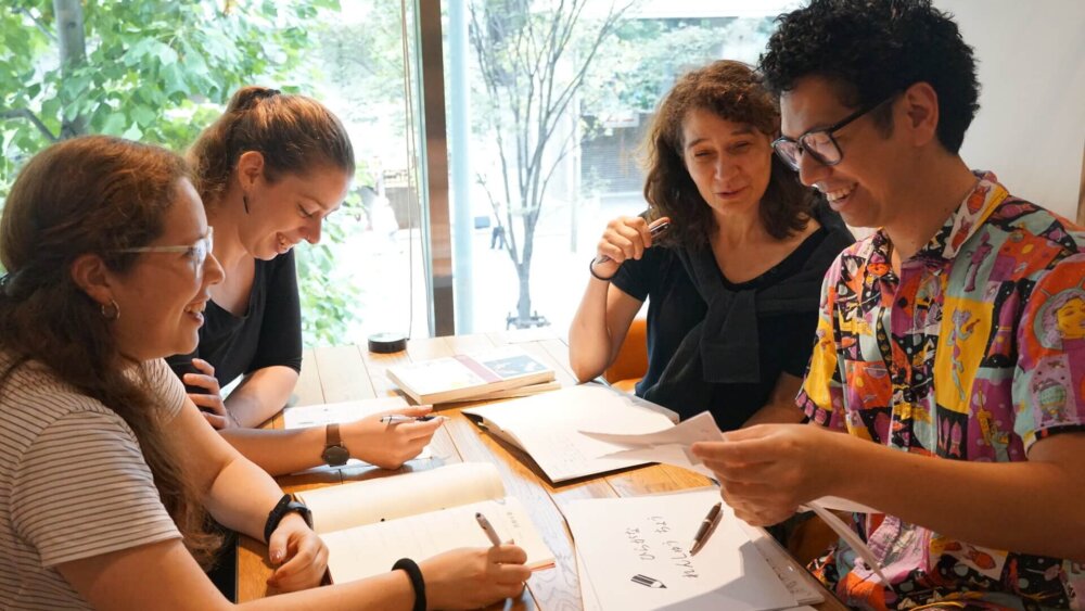 Vier junge Erwachsene lernen gemeinsam an einem Holztisch in einem hellen Raum mit großen Fenstern in Japan.