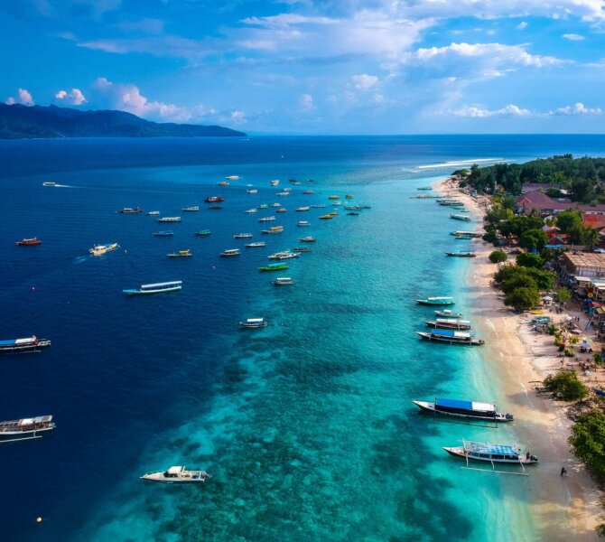 Luftaufnahme der Küste von Lombok mit türkisblauem Meer, vielen Booten und Strand mit Gebäuden und Palmen bei sonnigem Wetter