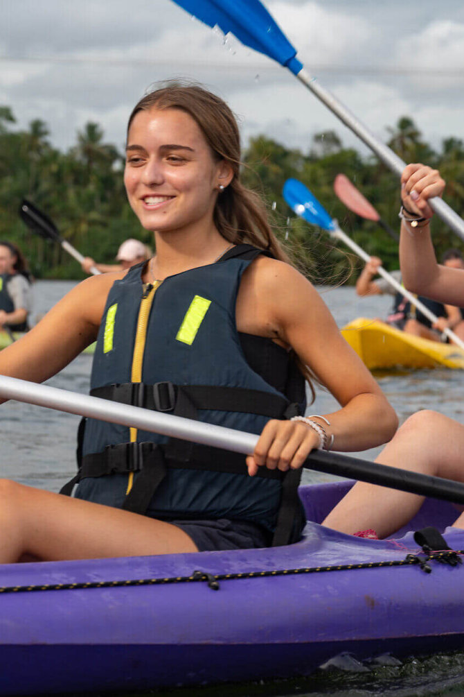 Zwei junge Frauen paddeln lachend im Kajak über einen tropischen Fluss, umgeben von Palmen und Natur auf Sri Lanka.