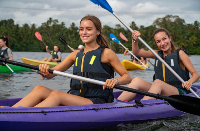 Zwei junge Frauen paddeln lachend im Kajak über einen tropischen Fluss, umgeben von Palmen und Natur auf Sri Lanka.