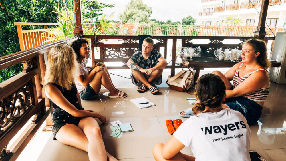 Gruppe junger Menschen sitzt auf einer Terrasse in tropischer Umgebung und tauscht sich während einer Way Changer Reise aus