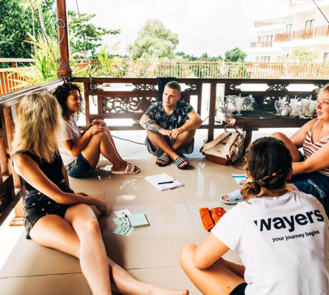 Gruppe junger Menschen sitzt auf einer Terrasse in tropischer Umgebung und tauscht sich während einer Way Changer Reise aus