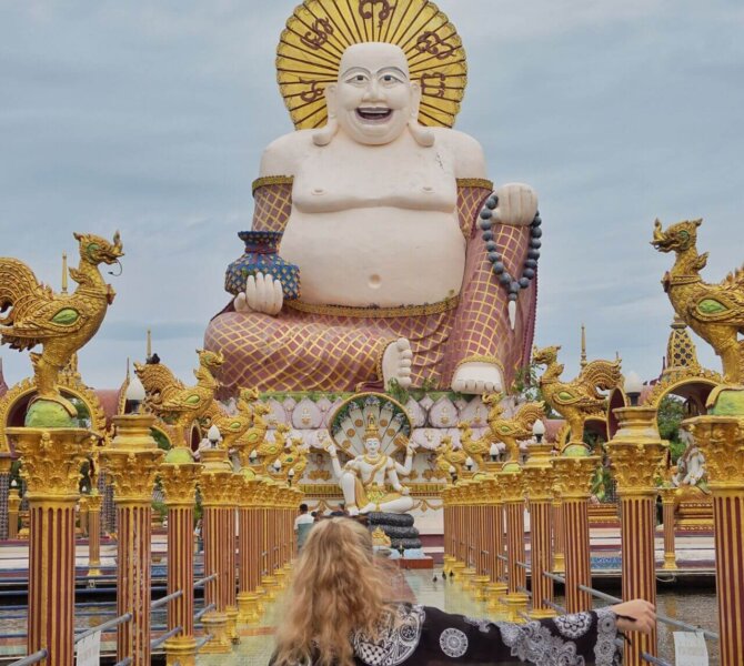 Person mit Tuch steht vor großer lachender Buddha-Statue auf kunstvoll verziertem Tempelgelände in Koh Samui unter leicht bewölktem Himmel