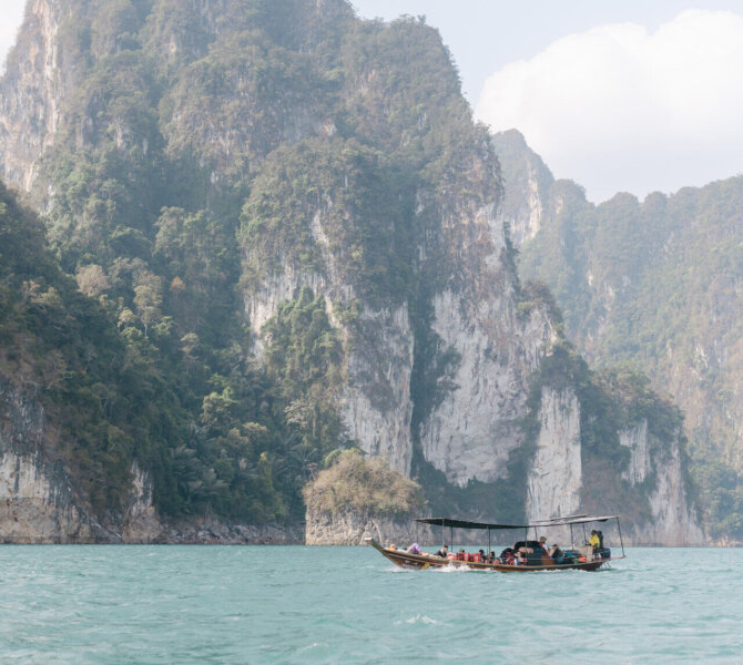 Langboot mit Menschen fährt über türkisfarbenes Wasser vor steilen grünen Kalksteinfelsen unter leicht bewölktem Himmel