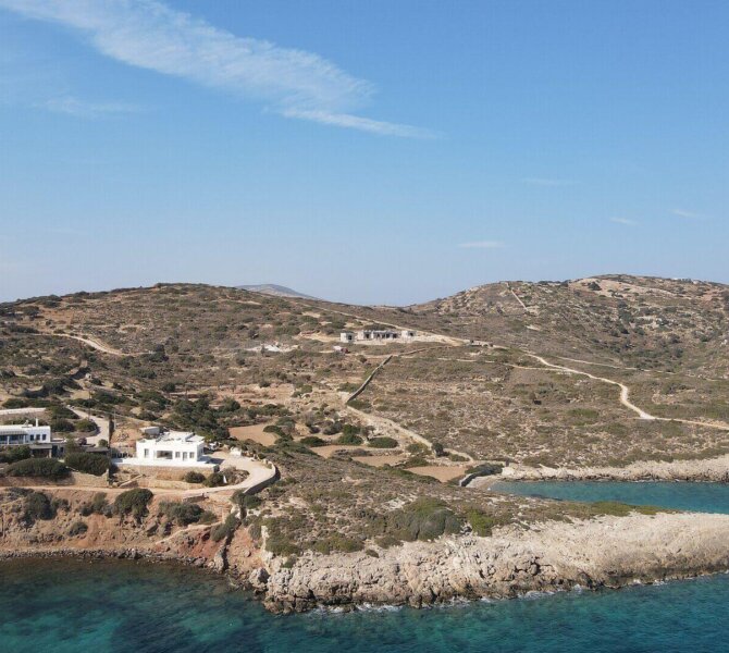Küste mit felsiger Landzunge und klarblauem Meer vor hügeliger Landschaft mit vereinzelten Häusern unter blauem Himmel