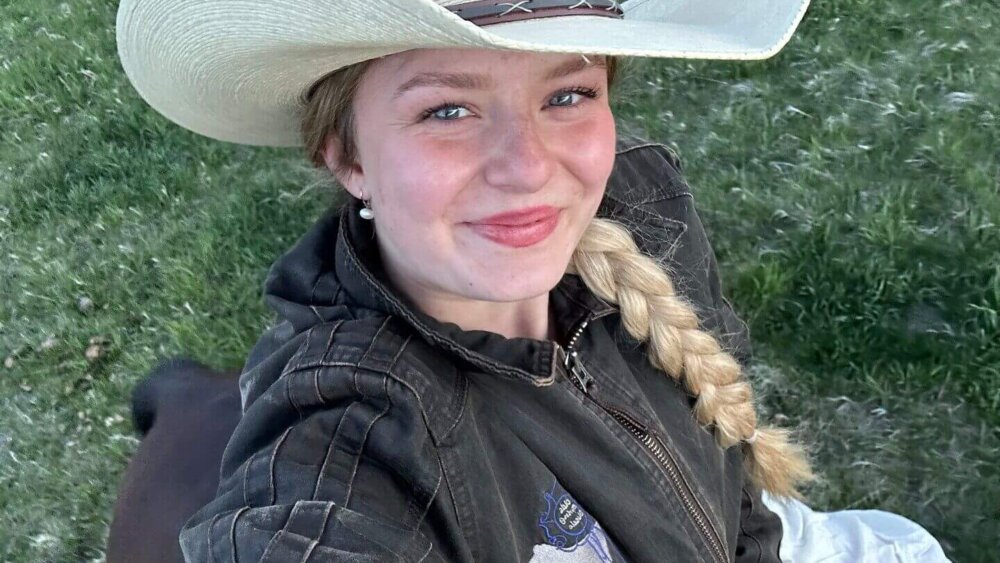 Junge Frau mit Cowboyhut und geflochtenem Zopf macht ein Selfie auf einem Pferd, im Hintergrund grüne Wiese.