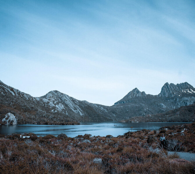 Blick auf einen ruhigen Bergsee mit umliegender Hügellandschaft und markanten Gipfeln unter blauem Himmel.