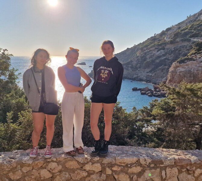 Drei junge Frauen posieren auf einer Steinmauer vor einer sonnigen Küstenlandschaft mit Meer Felsen und Pinien.