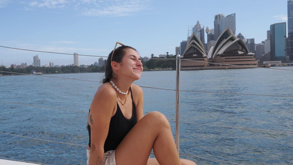 Junge Frau sitzt entspannt auf einem Boot vor dem Opernhaus in Sydney mit Blick auf das Wasser und die Skyline.