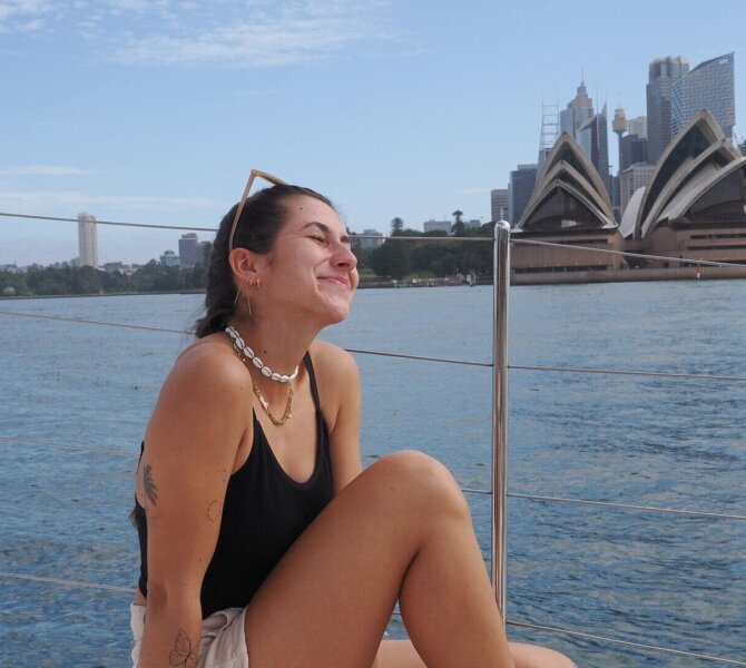 Junge Frau sitzt entspannt auf einem Boot vor dem Opernhaus in Sydney mit Blick auf das Wasser und die Skyline.