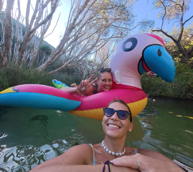 Zwei Frauen genießen das Wasser, eine auf einem bunten Papagei-Schwimmreifen, die andere lachend im Vordergrund mit Sonnenbrille.