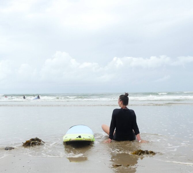 Frau sitzt im Neoprenanzug am Strand mit Blick aufs Meer, neben ihr ein Surfbrett im flachen Wasser.