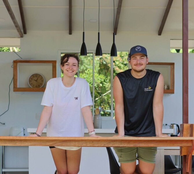Zwei junge Erwachsene lehnen lächelnd an einer Holztheke in einem offenen, modernen Küchenbereich mit Blick ins Grüne.