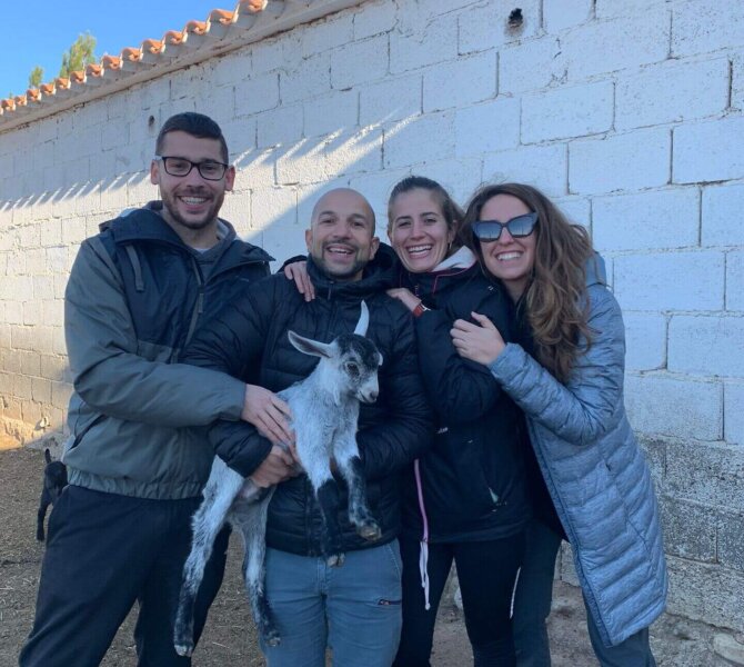 Vier lachende Personen posieren vor einer weißen Mauer, eine von ihnen hält eine kleine Ziege im Arm.
