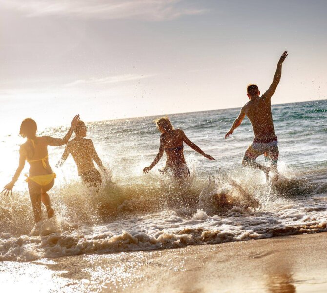 Vier Personen toben lachend in der Brandung am Strand bei Sonnenuntergang.