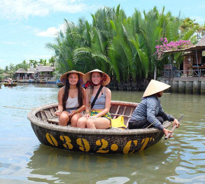 Zwei Touristinnen sitzen in einem traditionellen vietnamesischen Rundboot, das von einer Person mit Strohhut auf einem Fluss gerudert wird.