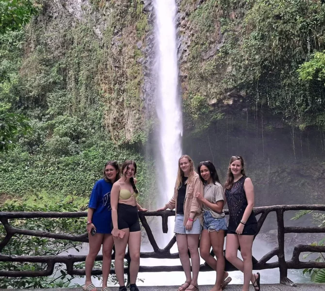 Fünf junge Frauen posieren lächelnd vor einem Wasserfall im tropischen Regenwald in Costa Rica.