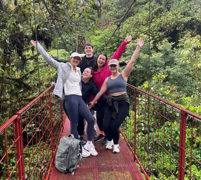Gruppe fröhlicher Menschen posiert auf einer roten Hängebrücke im Nebelwald von Costa Rica, umgeben von dichtem Grün.