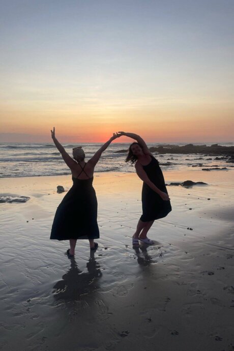 Zwei Frauen tanzen barfuß am Strand von Costa Rica bei Sonnenuntergang, das Meer und die Felsen im Hintergrund.