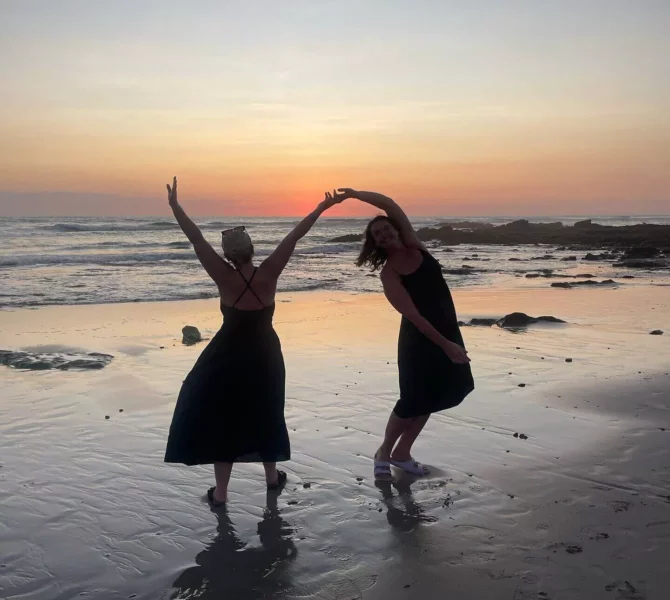 Zwei Frauen tanzen barfuß am Strand von Costa Rica bei Sonnenuntergang, das Meer und die Felsen im Hintergrund.