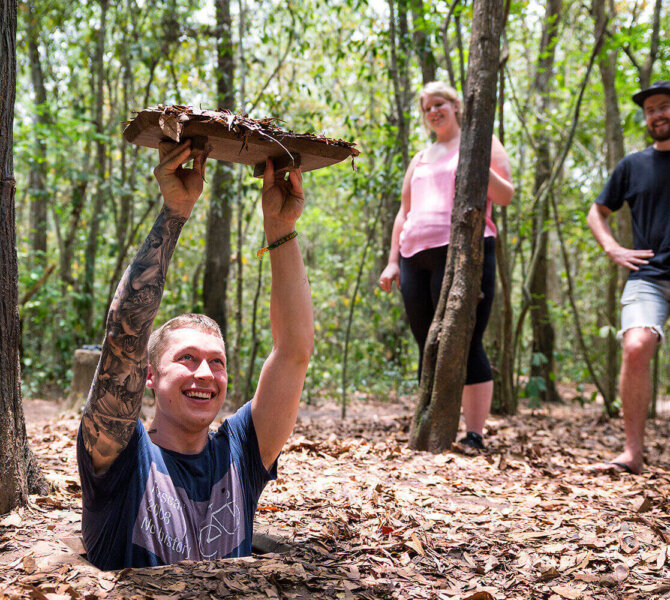 Ein Tourist steigt lachend aus einem schmalen Eingang der Cu-Chi-Tunnel, während zwei weitere Personen im Hintergrund zusehen.