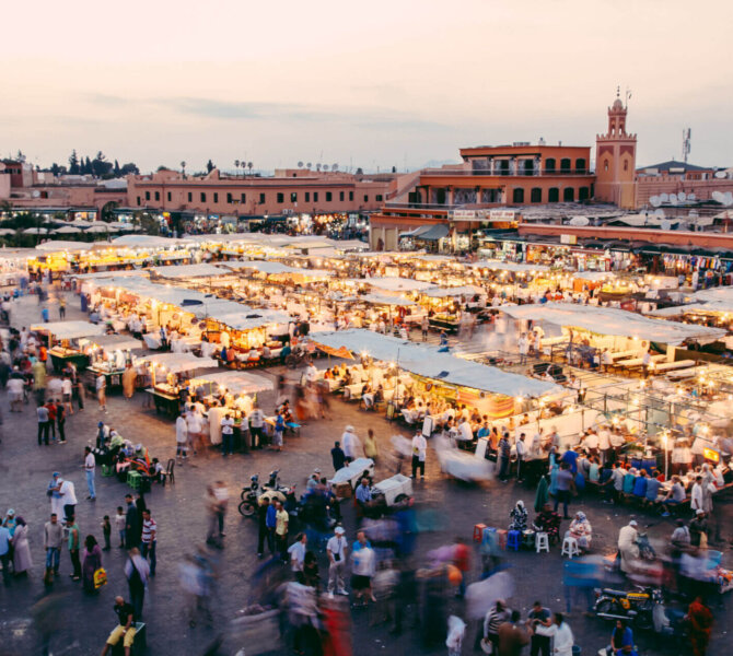 Belebter Marktplatz in Marrakesch bei Sonnenuntergang mit zahlreichen Ständen, Menschenmengen und Blick auf die Koutoubia-Moschee im Hintergrund.