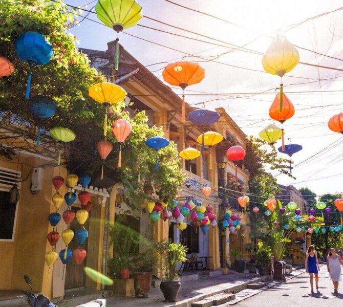 Bunte Laternen hängen über einer sonnigen Straße in Hoi An, Vietnam, während Touristinnen durch die charmante Altstadt schlendern.
