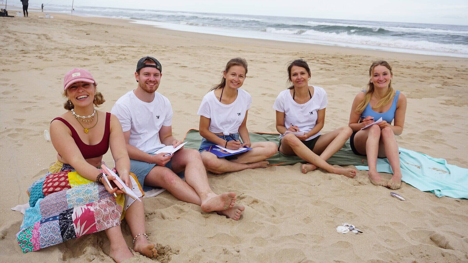 Fünf junge Erwachsene sitzen lachend im Sand am Strand und schreiben in Hefte während das Meer im Hintergrund rauscht.