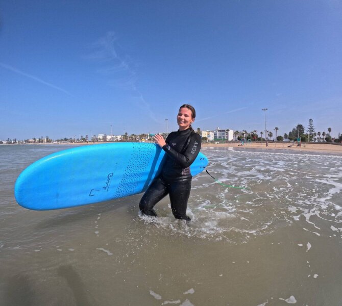 Surferin im Neoprenanzug steht lächelnd mit einem blauen Surfbrett im seichten Wasser vor der Küste von Essaouira, Marokko, bei klarem Himmel.