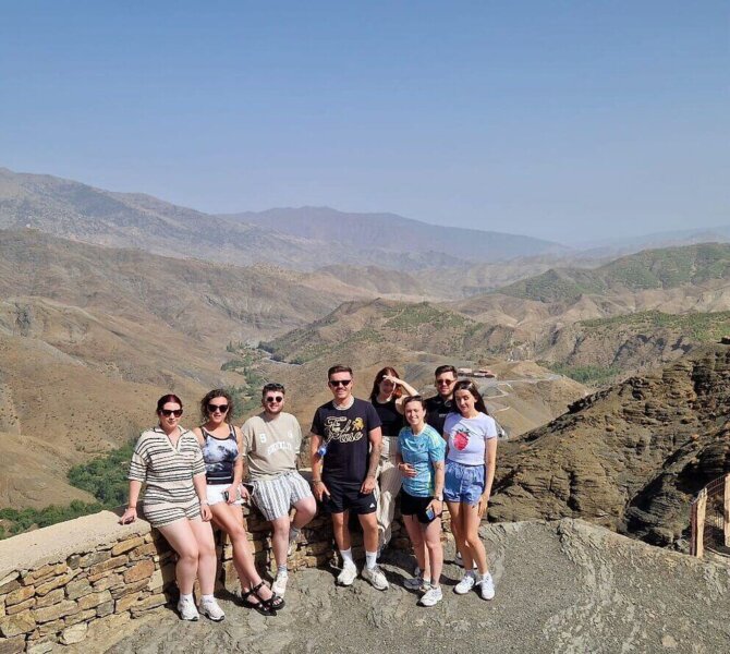 Gruppe von Touristen posiert vor beeindruckender Bergkulisse im Atlasgebirge in Marokko bei klarem, sonnigem Wetter.