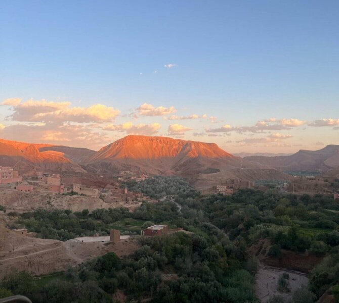 Landschaftsaufnahme eines grünen Tals im Dades Valley bei Sonnenuntergang, mit rötlich beleuchteten Bergen und klarem Himmel im Hintergrund.