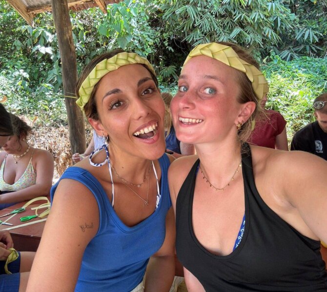 Zwei Frauen mit geflochtenen Stirnbändern lächeln in die Kamera, im Hintergrund basteln andere inmitten tropischer Vegetation.