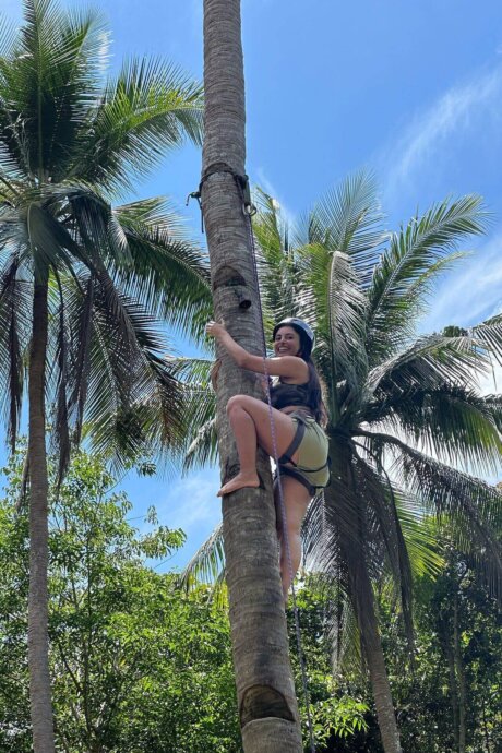 Frau klettert barfuß mit Sicherung an einer Palme empor, umgeben von tropischen Bäumen und blauem Himmel auf den Philippinen.