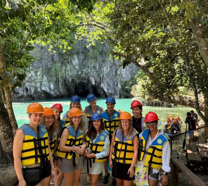Gruppe mit Helmen und Schwimmwesten vor einer Höhle am türkisfarbenen Fluss im tropischen Wald auf den Philippinen.