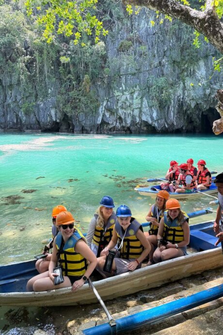 Gruppe junger Menschen mit Helmen und Schwimmwesten sitzt in einem Boot auf türkisblauem Wasser vor Felskulisse auf den Philippinen.