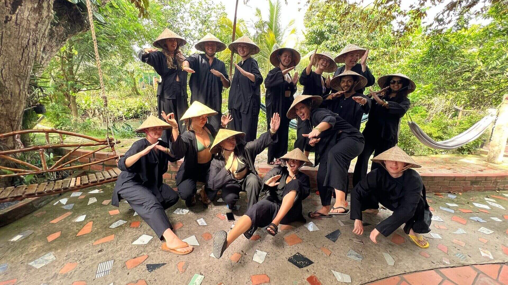 Gruppe junger Menschen in dunkler Kleidung und Strohhüten posiert gemeinsam vor grüner, tropischer Kulisse in Vietnam.