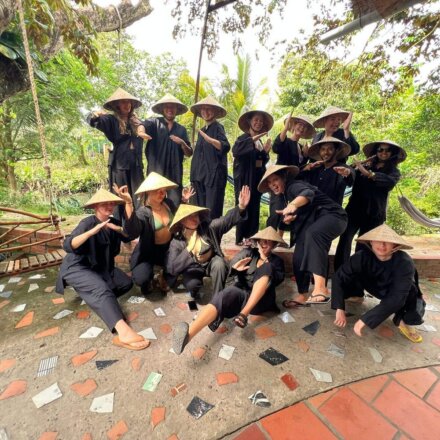 Gruppe junger Menschen in dunkler Kleidung und Strohhüten posiert gemeinsam vor grüner, tropischer Kulisse in Vietnam.