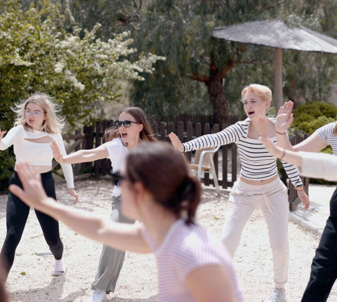 Gruppe von Frauen nimmt im Freien an einer Bewegungs- oder Achtsamkeitsübung teil, mit erhobenen Armen, umgeben von Bäumen und Sonne.