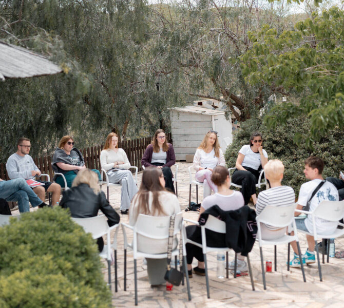 Gruppe von Menschen sitzt in einem Stuhlkreis im Freien unter Bäumen, vermutlich bei einem Workshop oder Gruppengespräch in entspannter Atmosphäre.