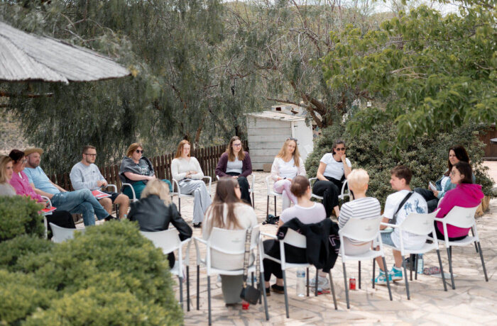 Gruppe von Menschen sitzt in einem Stuhlkreis im Freien unter Bäumen, vermutlich bei einem Workshop oder Gruppengespräch in entspannter Atmosphäre.