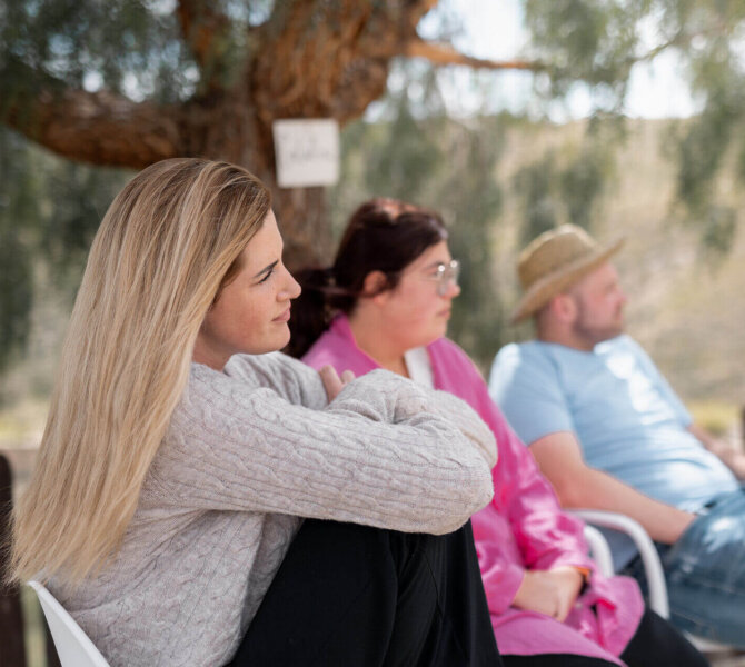 Mehrere Personen sitzen entspannt unter einem Baum im Schatten und lauschen aufmerksam, im Hintergrund eine ruhige ländliche Landschaft.