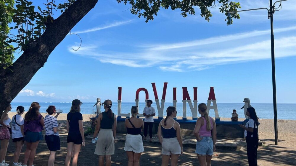 Eine Gruppe von jungen Menschen steht im Gegenlicht vor einem Schild, wo in großen Buchstaben LOVINA steht. Im Hintergrund ist der Strand und das Meer.
