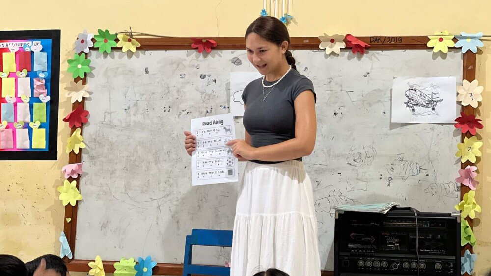 Ein Mädchen steht vor einer Wand mit Tafel und hält ein Arbeitsblatt in der Hand und präsentiert es den Kindern, die vor ihr sitzen. Von den Kindern sieht man nur die Hinterköpfe.