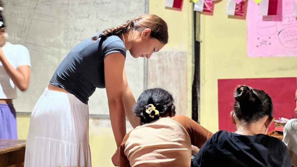 Ein Mädchen in T-Shirt und weißem Rock beugt sich von der Seite über einen Schreibtisch an dem zwei Kinder sitzen, die von hinten zu sehen sind.
