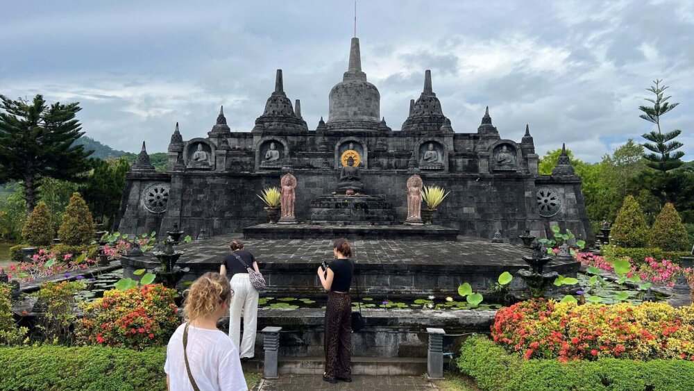 Ein paar Menschen laufen durch eine Gartenanlage in der zentral ein Tempel steht. Der Himmel ist bewölkt und die Blumen und Büsche an den Seiten farbenfroh.