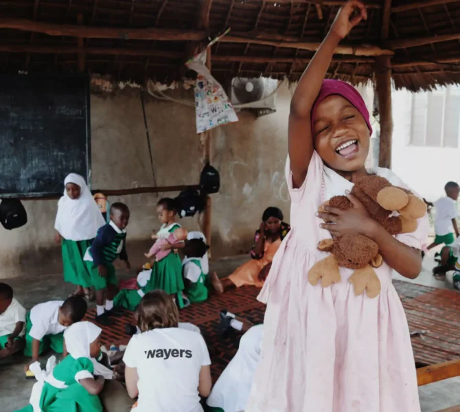 Fröhliches Mädchen in rosa Kleid mit Stofftier hebt lachend den Arm in einer Schule mit Kindern und wayers Freiwilligen