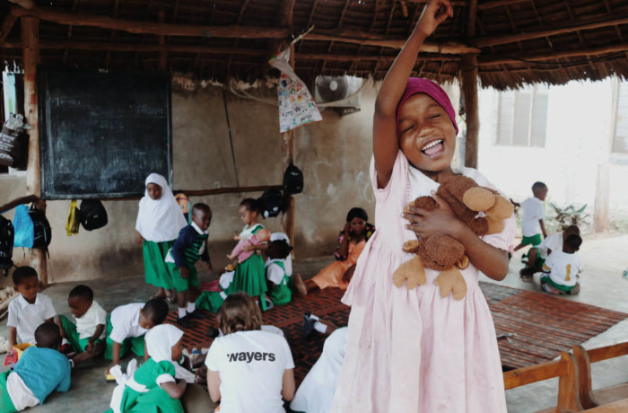 Fröhliches Mädchen in rosa Kleid mit Stofftier hebt lachend den Arm in einer Schule mit Kindern und wayers Freiwilligen