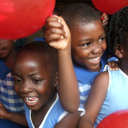 Lachende Kinder mit roten Luftballons in Schuluniform bei fröhlicher Aktivität im Rahmen von Sozialarbeit mit Kindern in Ghana
