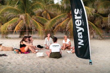 Teilenehmer sitzen im Sand vor Palmen neben einem schwarzen wayers Banner und nehmen an einem Coaching teil
